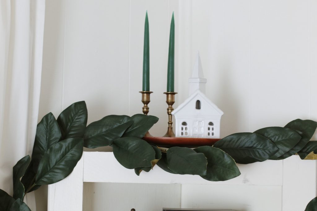 white ceramic church and 2 brass candlesticks on cabinet with greenery